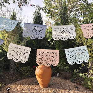 May include: A string of colorful paper banners with cut-out designs hangs between trees in a garden setting. A terracotta pot sits in the center of the image.