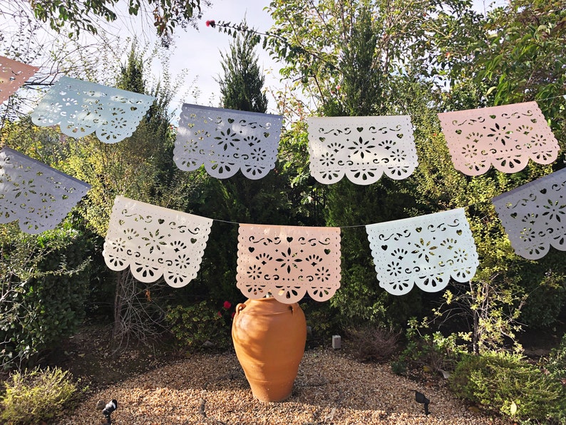 May include: A string of colorful paper banners with cut-out designs hangs between trees in a garden setting. A terracotta pot sits in the center of the image.