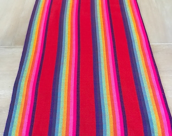 May include: A colorful table runner with a rainbow stripe pattern and fringe edges. The runner is made of woven fabric and has a red background with stripes of pink, orange, yellow, green, blue, and purple.