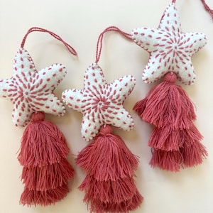 May include: A garland of white felt star ornaments with red embroidered details and matching tiered tassels. Each star has a string for hanging. The tassels are a deep rose color, adding a touch of elegance to the festive decor.