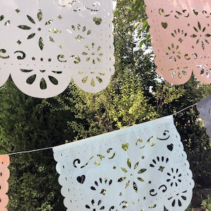 May include: A string of paper cutouts, or "papel picado", in white, light blue, pink, and gray, hanging against a backdrop of green foliage. The cutouts feature intricate designs of flowers, hearts, and swirls.