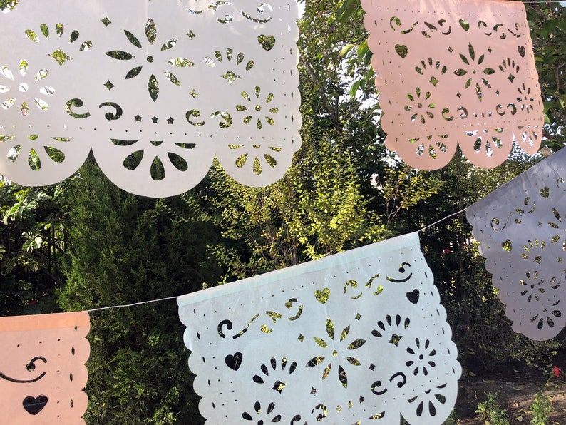 May include: A string of paper cutouts, or "papel picado", in white, light blue, pink, and gray, hanging against a backdrop of green foliage. The cutouts feature intricate designs of flowers, hearts, and swirls.