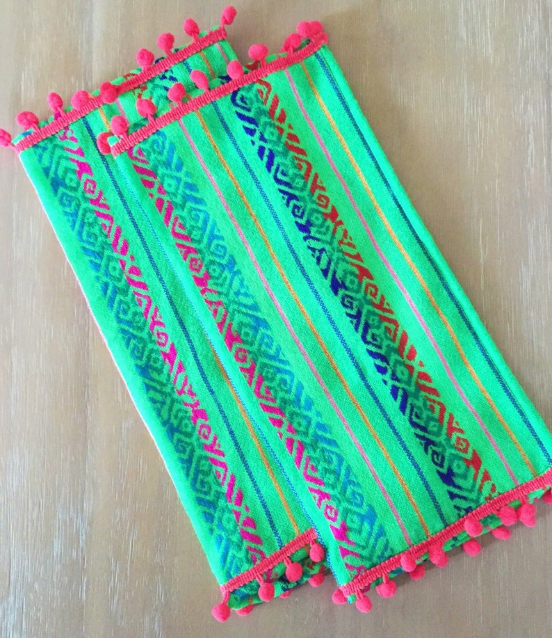 May include: Two green tea towels with a colorful geometric pattern and a pink pom-pom trim.