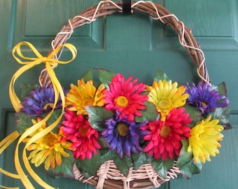 May include: A wreath made of woven twigs with a yellow ribbon bow and a cluster of colorful artificial flowers. The flowers are red, yellow, purple, and green.