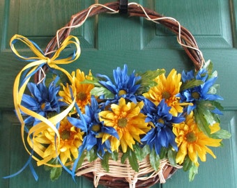 May include: A decorative wreath featuring a woven vine base, adorned with vibrant yellow and blue artificial flowers. A woven basket shape holds the flowers, and a yellow and blue ribbon bow adds a finishing touch. The wreath hangs on a green door.