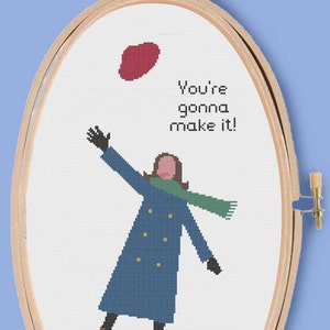 May include: Cross-stitch embroidery in a wooden hoop featuring a woman in a blue coat and green scarf reaching up to catch a red heart with the text "You're gonna make it!"