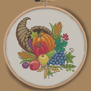 Puede incluir: Un diseño de bordado de punto de cruz que presenta una cornucopia desbordante de frutas y verduras de la cosecha de otoño, incluyendo una calabaza, uvas, manzanas, maíz y un pimiento verde. La cornucopia es marrón y tiene un ribete dorado.