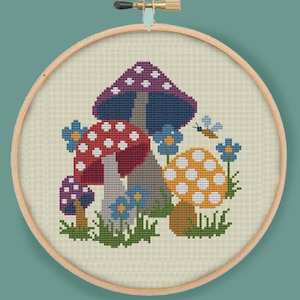 Könnte beinhalten: Stickrahmen mit einer Kreuzstich-Stickerei, die eine fantasievolle Szene mit bunten Pilzen mit weißen Punkten, blauen Blumen und einem kleinen fliegenden Insekt zeigt. Die Pilze sind in einer Gruppe angeordnet, wobei der größte Pilz hinten und der kleinste Pilz vorne ist. Der Hintergrund ist ein hellbeiger Stoff.