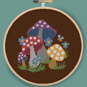 Könnte beinhalten: Ein Kreuzstich-Stickrahmen mit braunem Hintergrund, auf dem drei Pilze mit weißen Punkten, lila, blauen und gelben Akzenten und blauen Blumen zu sehen sind.