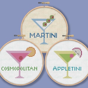 May include: Three embroidery hoops with cross-stitch designs of cocktails. The top hoop has a blue martini with olives and the text "MARTINI". The left hoop has a pink cosmopolitan with a cherry and the text "COSMOPOLITAN". The right hoop has a green appletini with a cherry and a lime wedge and the text "APPLETINI".