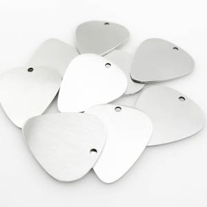 May include: A group of ten silver-colored metal guitar picks with a hole at the top. The picks are arranged in a pile on a white surface.