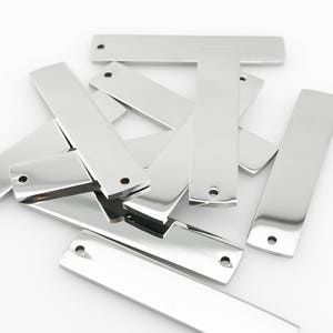 May include: A collection of shiny, rectangular metal tags with rounded edges and a small hole at one end. The tags are silver in color and appear to be made of stainless steel. They are scattered on a white surface.