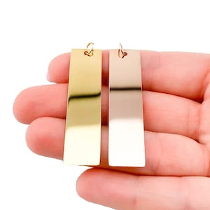 Puede incluir: Dos etiquetas rectangulares de metal con un agujero en la parte superior. La etiqueta de la izquierda es dorada y la etiqueta de la derecha es plateada.