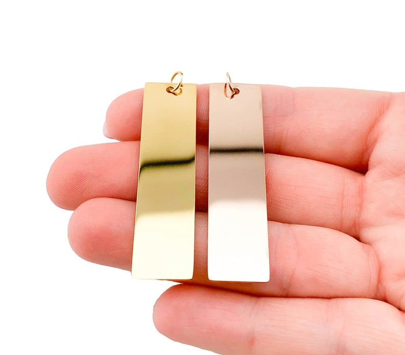 Puede incluir: Dos etiquetas rectangulares de metal con un agujero en la parte superior. La etiqueta de la izquierda es dorada y la etiqueta de la derecha es plateada.