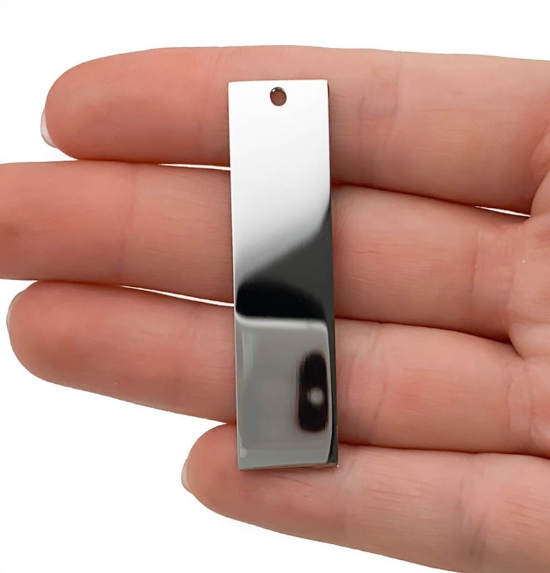 Puede incluir: Una etiqueta rectangular de metal plateado con un agujero en la parte superior. La etiqueta es lisa y brillante.