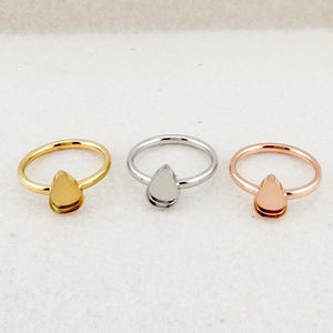 May include: Three rings with teardrop-shaped settings. One ring is gold, one is silver, and the other is rose gold. The rings are displayed on a light-colored surface. The rings are likely for jewelry making.