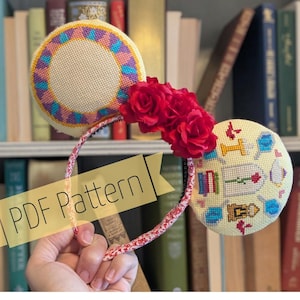 May include: A headband featuring two circular ears with cross-stitch embroidery. One ear has a geometric pattern in yellow, blue, and purple. The other ear displays small cross-stitched icons including a rose, books, and a clock. Three red fabric roses sit between the ears. A banner across the centre reads PDF Pattern.