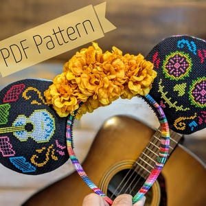 May include: A headband featuring two black circular ears with cross-stitch embroidery, one displaying a light blue guitar and the other a colorful skull design. A row of yellow marigold flowers sits between the ears. The band has a striped pattern.