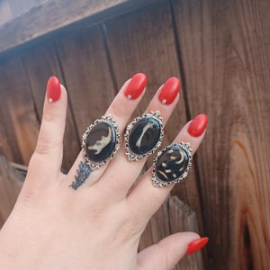 May include: Three silver-toned rings featuring oval settings with black resin centers. Each ring contains a small, light-colored bone fragment embedded within the resin. The settings have an ornate, decorative metal border around the oval shape.