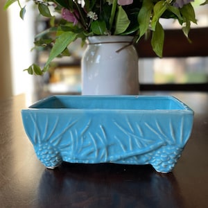 May include: A rectangular, turquoise ceramic planter with embossed pinecone and leaf designs. The planter sits on small feet and is filled with a bouquet of pink and purple flowers in a white vase.