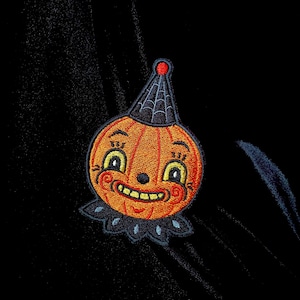 May include: A black fabric background with an orange and black embroidered pumpkin patch applique. The pumpkin has a smiling face with a black spider web on its hat.