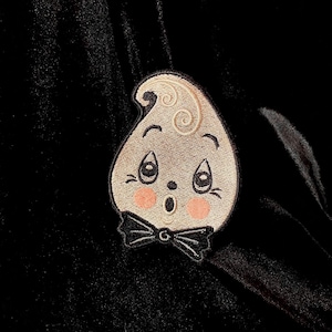 Puede incluir: Una tela de terciopelo negro con un fantasma blanco bordado con una pajarita negra. El fantasma tiene una expresión de sorpresa con ojos grandes y una boca pequeña.