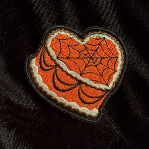 May include: Embroidered patch in the shape of a heart with an orange spiderweb design. The patch has a black border and is set against a black background. The heart shape is filled with an orange color.