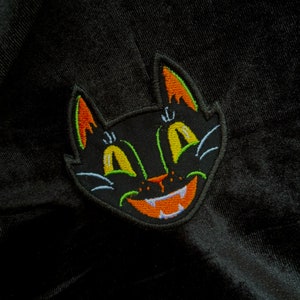 May include: Black embroidered patch of a cat face with orange and yellow details. The cat has a wide grin with sharp teeth and large eyes.