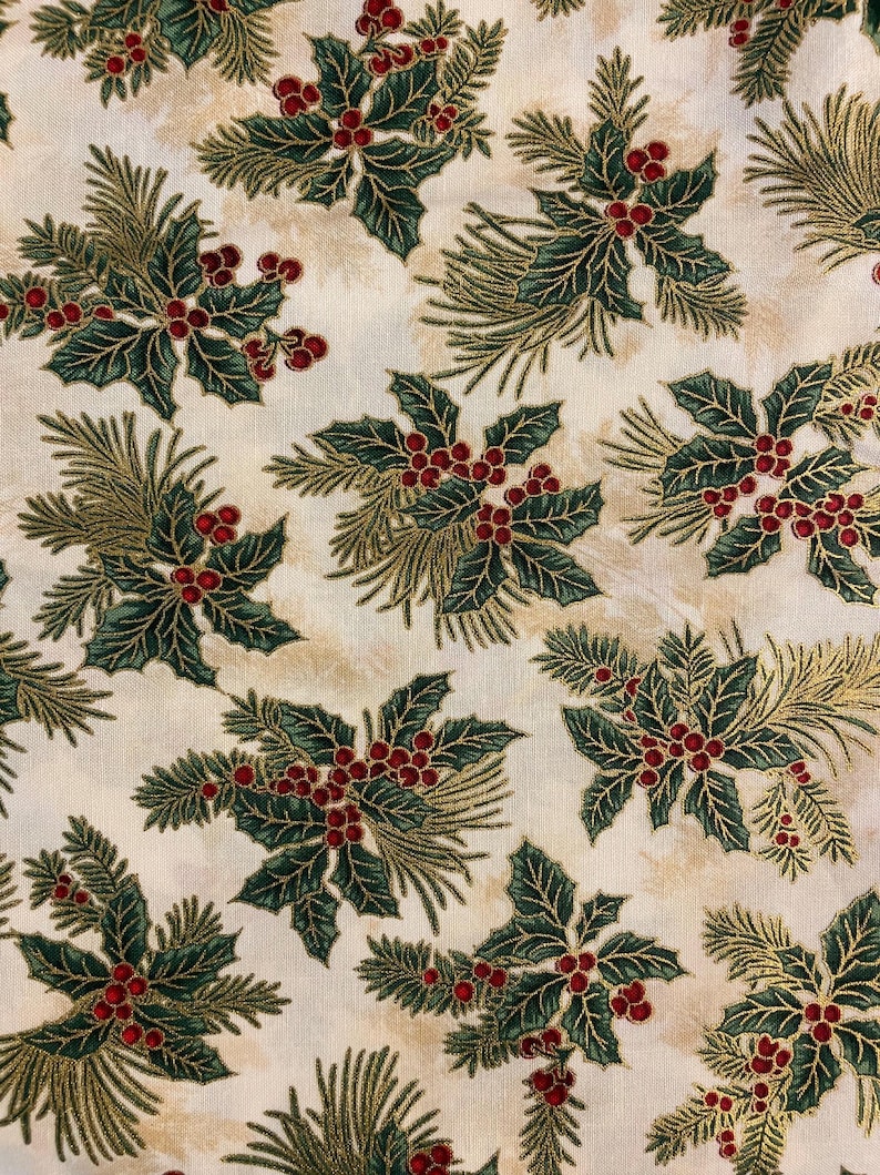 May include: A cream-coloured fabric featuring a repeating pattern of dark green holly leaves, clusters of small red berries, and sprigs of pine needles. The background has a subtle, mottled beige texture throughout the entire textile surface.