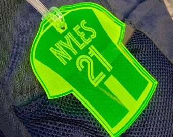 Op de afbeelding: Een felgroen bagagelabel in de vorm van een shirt, met de naam "NYLES" en het nummer "21" in het wit. Het label is bevestigd aan een doorzichtige plastic lus en hangt aan een donkerblauwe rugzak.