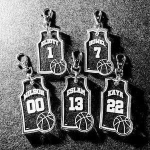 May include: Black and white basketball jersey keychains. Each keychain features a jersey design with a name, number, and a basketball. The jerseys are attached to silver key rings. The names include Elliott, Howell, Wilson, Eslam, and Kaya.