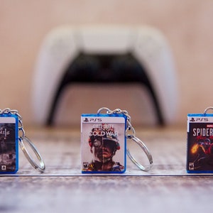 May include: Three blue and silver keychains with video game covers. The keychains feature the game covers for "Demon's Souls", "Call of Duty: Black Ops Cold War", and "Spider-Man: Miles Morales".