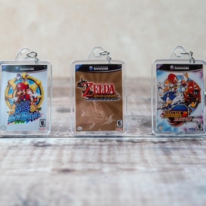May include: Three clear plastic keychains with colourful game cartridge designs. The keychains feature the games "Super Mario Sunshine", "The Legend of Zelda: The Wind Waker", and "Sonic Adventure 2: Battle".