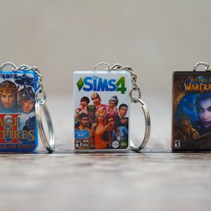 Könnte beinhalten: Drei Miniatur-Schlüsselanhänger in Form von Spielboxen. Der erste Schlüsselanhänger ist blau und trägt den Text "Age of Empires II The Age of Kings". Der zweite Schlüsselanhänger ist weiß und trägt den Text "Die Sims 4". Der dritte Schlüsselanhänger ist braun und trägt den Text "World of Warcraft".