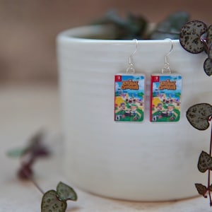 May include: A pair of silver dangle earrings with miniature Animal Crossing game cartridge charms. The charms feature the game's logo and a colourful illustration of characters on a beach.