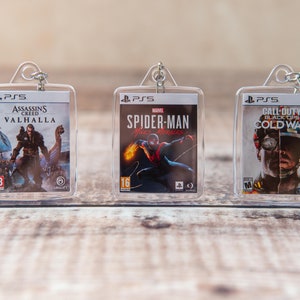 May include: Three clear plastic keychains with images of video game covers. The first keychain features the cover art for Assassin's Creed Valhalla, the second keychain features the cover art for Spider-Man: Miles Morales, and the third keychain features the cover art for Call of Duty: Black Ops Cold War.