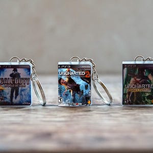 Könnte beinhalten: Drei Miniatur-Schlüsselanhänger mit rechteckigen Kunststoffgehäusen und aufgedruckten Covern für die PlayStation 3-Videospiele Call of Duty World at War, Uncharted 2 Among Thieves und Uncharted Drakes Fortune, jeweils an einem silbernen Metallring befestigt.