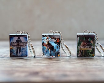 May include: Three miniature keychains featuring rectangular plastic cases with printed covers for the PlayStation 3 video games Call of Duty World at War, Uncharted 2 Among Thieves, and Uncharted Drakes Fortune, each attached to a silver metal ring.