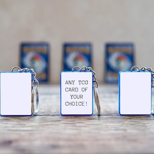Könnte beinhalten: Drei Schlüsselanhänger mit einem weißen rechteckigen Einsatz und einem blauen Rand. Der mittlere Schlüsselanhänger trägt den Text "ANY TCG CARD OF YOUR CHOICE!". Die Schlüsselanhänger liegen auf einer Holzoberfläche, mit verschwommenen Sammelkartenbildern im Hintergrund.
