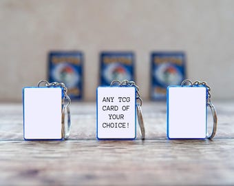 May include: Three rectangular blue-framed keychains with silver metal rings. The center keychain features the white text ANY TCG CARD OF YOUR CHOICE! on a white background, while the two flanking keychains have blank white centers.