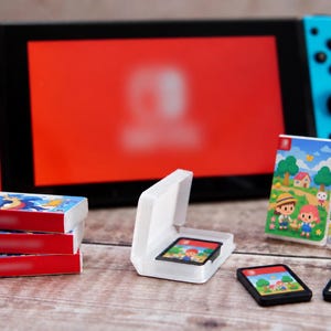 Op de afbeelding: Een draagbare videogameconsole met een rode linkercontroller en een blauwe rechtercontroller, vergezeld van verschillende miniatuurspelcartridges en twee kleine speldoosjes met kleurrijke cartoonillustraties.