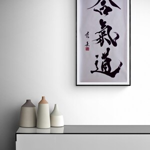 Puede incluir: Una impresi&oacute;n enmarcada en negro con fondo blanco y caligraf&iacute;a negra en caracteres chinos. La impresi&oacute;n est&aacute; colgada en una pared blanca sobre un armario blanco con tres jarrones de cer&aacute;mica en la parte superior.