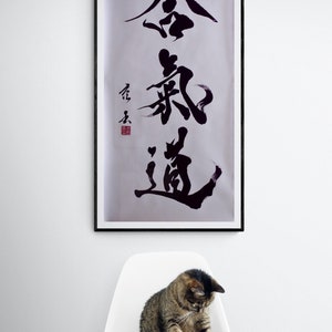 Puede incluir: Una obra de arte de caligraf&iacute;a en blanco y negro enmarcada con los caracteres "金 鍼 道" en un estilo japon&eacute;s tradicional. La obra est&aacute; colgada en una pared blanca sobre una silla blanca con un gato atigrado sentado en ella.