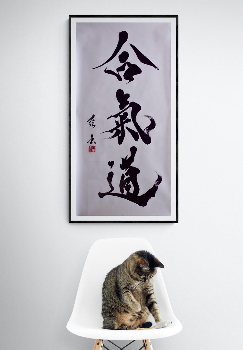 Puede incluir: Una obra de arte de caligraf&iacute;a en blanco y negro enmarcada con los caracteres "金 鍼 道" en un estilo japon&eacute;s tradicional. La obra est&aacute; colgada en una pared blanca sobre una silla blanca con un gato atigrado sentado en ella.