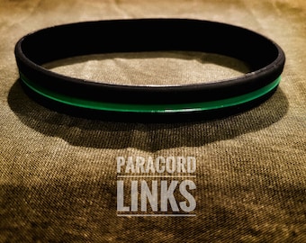 May include: Black paracord bracelet with a thin green stripe. The bracelet is on a brown fabric background. The text "PARACORD LINKS" is printed below the bracelet.