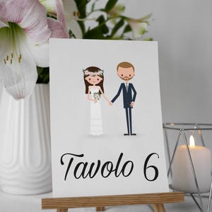 Può includere: Un segnaposto per il tavolo nuziale con un'illustrazione a cartoni animati di sposi che si tengono per mano. La carta è esposta su un piccolo cavalletto di legno. Il testo "Tavolo 6" è scritto in elegante corsivo.