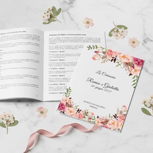 Può includere: Libro aperto e invito di nozze con design floreale. L'invito recita "La Cerimonia Romeo e Giulietta 20 giugno 2027". Nastro rosa e fiori sparsi su una superficie di marmo.