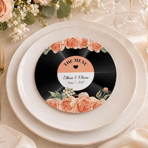 Può includere: Un menu circolare progettato per assomigliare a un disco in vinile nero, con un'etichetta centrale che riporta il testo THE MENU, un'icona a forma di cuore, Ethan & Olivia e June 7, 2027. Il disco è decorato con rose color pesca e foglie verdi.