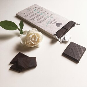 May include: A partially unwrapped bar of dark chocolate, with the brand name "Lindt" embossed on the pieces. A white rose and green leaves are next to the chocolate. The packaging has text and a heart symbol.