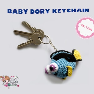Op de afbeelding: Een gehaakte Baby Dory sleutelhanger in blauw, zwart en geel, bevestigd aan een sleutelring met drie sleutels. De sleutelhanger heeft grote, gedetailleerde ogen en een vinnenontwerp. De tekst bovenaan luidt "BABY DORY KEYCHAIN".